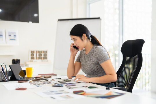 Concentrated young interior designer woman sitting at table with blueprint drawing ,swatches and material , discussing interior design with client by phone, choosing materials for decorations.