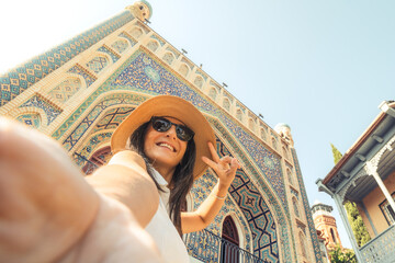 Smiling woman takes selfie at camera show peace sign by colorful Persian-style mosaic architecture in Abanotubani district during sunny summer vacation in Tbilisi, Georgia – concept travel freedom joy