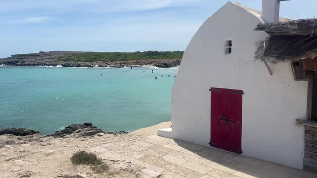 A charming beachside scene at Binibeca Nou with a traditional whitewashed building, a red door, and turquoise waters in the background. People are enjoying the water, capturing a tranquil atmosphere.