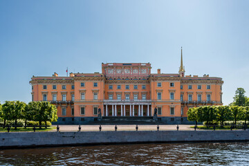 Mikhailovsky Castle of pink color with columns and a spire, stands on the river bank under a clear sky.