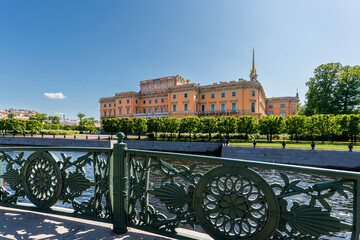 Mikhailovsky Castle is visible behind an openwork bridge fence and a river under a blue sky.