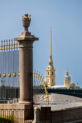A view of the Peter and Paul Cathedral spire and a bridge over the river, framed by a forged grille and column.
