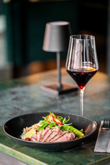 A gourmet dish with sliced duck breast, fresh greens, and grains, elegantly plated in a black bowl. A glass of red wine and a table lamp create a stylish fine-dining atmosphere.