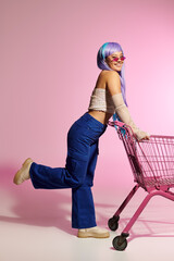 Stylish woman with vibrant hair poses playfully with shopping cart against pink backdrop