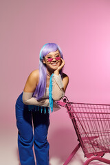 Stylish woman posing with colorful wig and shopping cart against a vibrant pink backdrop