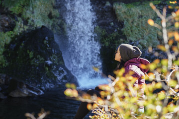 Woman in a beanie and winter coat sits contemplatively on rocks near the base of a waterfall, surrounded by autumn foliage