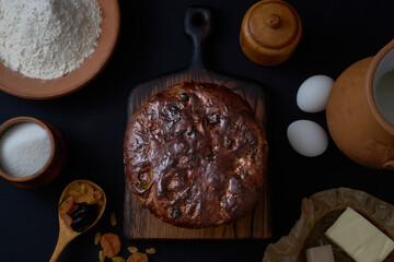 Easter cake rises on a wooden board surrounded by flour, sugar, milk, eggs, butter, and dried fruits on a black background