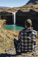 Man in a plaid shirt and cap sits on a rocky outcrop, overlooking Salto del Agrio waterfall. Neuquen, Argentina