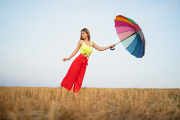 A young woman enjoys a sunny day in a vibrant field. She holds a colorful umbrella, adding charm and energy to the picturesque rural scene. Her cheerful outfit complements the lively atmosphere.
