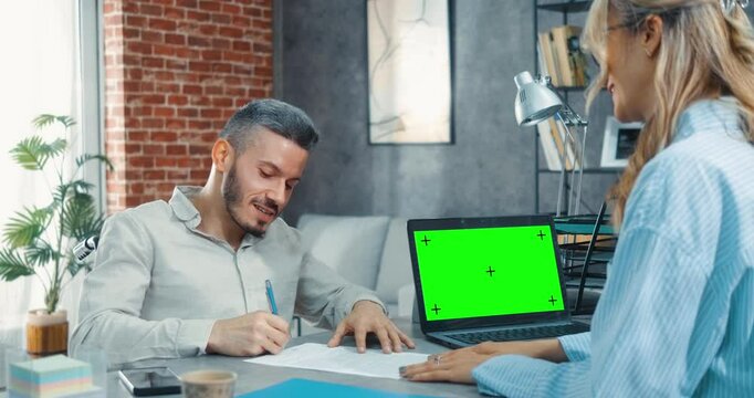 Laptop with mock-up green screen chromakey display on desk while a satisfied man in a wheelchair signs employment contract during job interview with female employer in modern office.