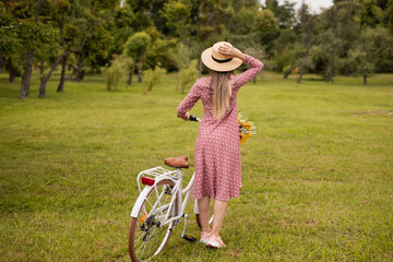 A woman in a polka dot dress and straw hat holding a bicycle in a green park. The scene conveys...