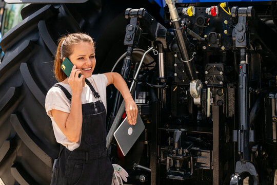 Female mechanic speaking on mobile phone next to tractor. - Powered by Adobe