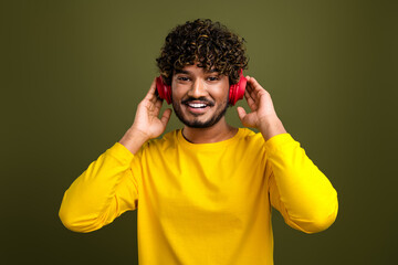 Young man in yellow shirt wears headphones and smiles joyfully against a green background for...