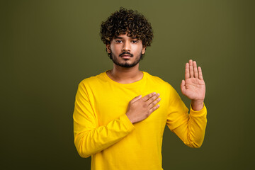 Young man in casual yellow shirt raising hand in affirmation while posing on green background with...