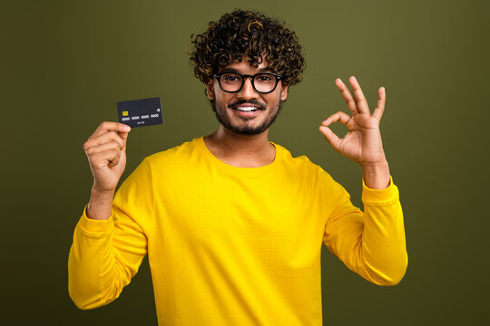 Confident young man in a casual yellow shirt holding a black credit card and gesturing okay sign against green background