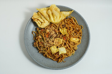 A plate of Indonesian spicy fried noodles served with crispy fried wontons and chili flakes, presented on a grey ceramic plate. Popular street food known for its rich and savory taste.