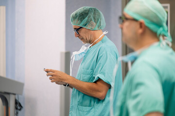 Side view of male doctor using smartphone in hospital corridor while wearing teal scrubs and green...