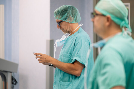 Side view of male doctor using smartphone in hospital corridor while wearing teal scrubs and green surgical cap