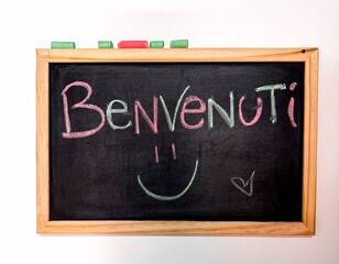 Obraz premium A warm welcome is displayed on a wooden chalkboard, featuring the word 'Benvenuti' in colorful chalk. A smiley face adds a cheerful touch in a cafe environment