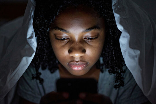 A young african american kid in a in bed under the blanket, her face lit by the light from her phone. The risks of cyber danger and the impact of social media platforms on teenagers and kids.