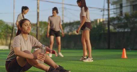 Indian happy athlete woman lady player group play football game kicking soccer ball enjoy team outdoor young adult gen z tired girl female sitting take break practice health care pick up drink water - Powered by Adobe