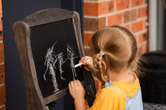 Little girl standing outside in backyard drawing her family on chalkboard