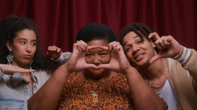Chest up of cheerful senior Black woman in leopard print blouse forming heart shape with her hands while sitting between her two smiling grandkids in cozy photo booth with red curtains