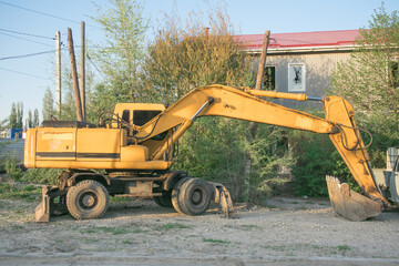 Excavator parked under clear blue sky near overgrown area and weathered building