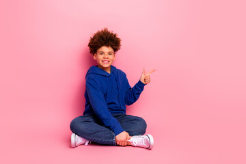 Happy boy sitting cross legged in blue hoodie on pink background, pointing in gesture, cheerful and...