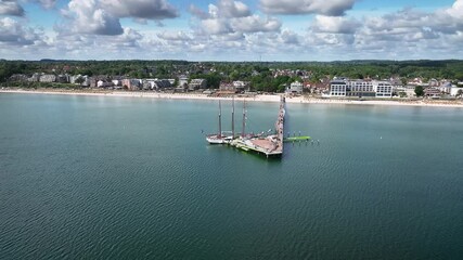 Aerial view of tall ship moored at long pier in Scharbeutz, coastal resort on the Baltic Sea in Schleswig-Holstein, Germany. The camera retreats and pans right to left, revealing seaside town. - Powered by Adobe
