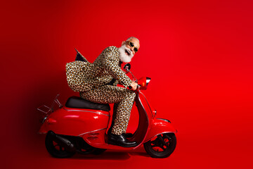Fashionable elderly man wearing leopard print suit rides red scooter against red background exuding humor and eccentric style
