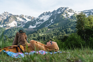 Man hiker relaxing on blanket in front of scenic snow-capped mountains and lush green valley, enjoying summer nature outdoors, in meadow with beautiful panoramic view of snowy peaks and forested hills