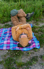 Top view of a man resting in yellow hoodie on a colorful checkered blanket on the ground, surrounded by grass and nature, hiking and nature exploration