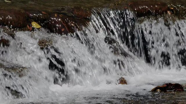 Panning slow motion view of small cascades of river water falling through an elevated stone ledge in sunny weather cerating foams and splashes.