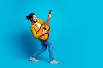 Cheerful young man with curly hair enthusiastically playing an acoustic guitar on a vibrant blue...