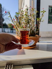 Hand holding traditional tulip-shaped glass of Turkish tea against sunlit courtyard. Antalya, Turkey, cultural tradition, tea time, warm light, outdoor scene, Mediterranean lifestyle, local culture. 