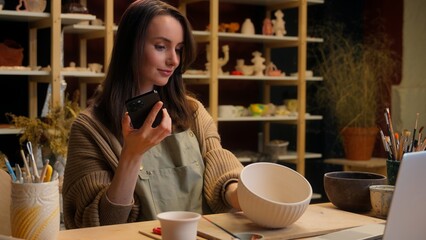 Woman blogger sculptor potter take photo mobile phone craft handmade bowl crockery use laptop Caucasian girl female artisan small business owner sale ceramics online shop crafting in pottery workshop