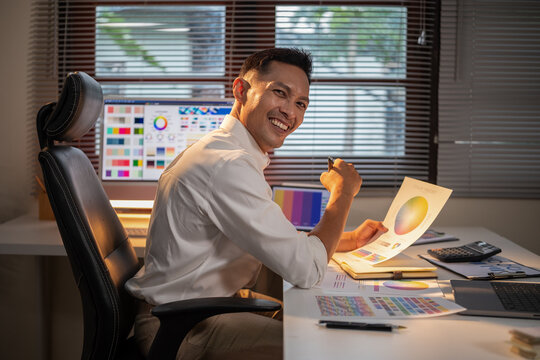 Smiling male graphic designer holding a color theory chart while working in a creative office space.