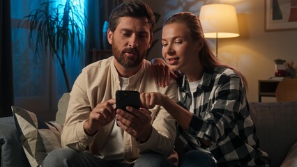 Caucasian family couple man woman home evening using smartphone together online shopping on sofa husband wife girl guy buying internet purchase choose goods home renovation night browsing mobile phone
