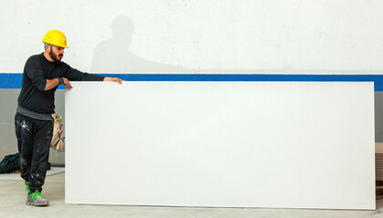 Construction worker holding large white plasterboard sheet in a construction site
