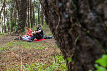 Two men in the forest resting and enjoying nature. Lying on a red blanket, reading, eating, and chatting
