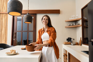 Young woman in an orange shirt and white pants smiles while holding a bowl in a modern kitchen,...