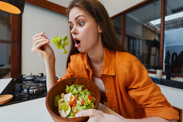 Surprised young woman with long hair in casual orange shirt, holding a bowl of fresh salad, showcasing healthy eating in a modern kitchen setting
