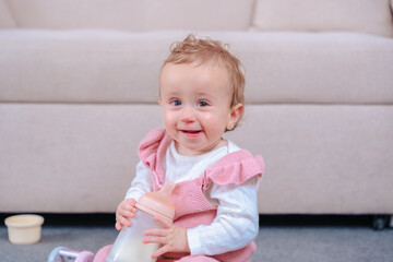 Fototapeta premium Adorable baby girl sits on the carpet holding a milk bottle, smiling in front of a beige sofa. Perfect for themes related to childhood, growth, nutrition, happiness, and home comfort. 