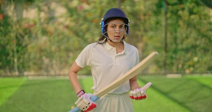 Indian gen z athlete sports woman lady standing at outdoor play ground wear white uniform and protective helmet looking at camera asian cricket player female girl hold bat day time at match playground