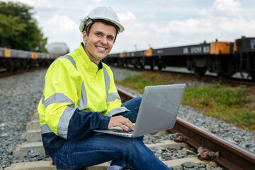 Smiling male railway engineer in reflective green work clothes and white helmet uses laptop computer while sitting on railway tracks and looking at camera