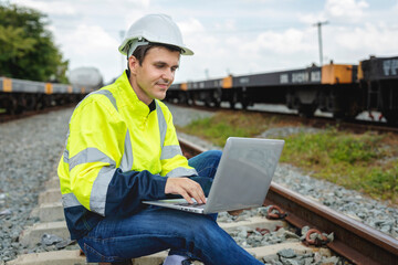 Male railway engineer in reflective green work clothes and white helmet uses laptop computer while sitting on railway tracks.