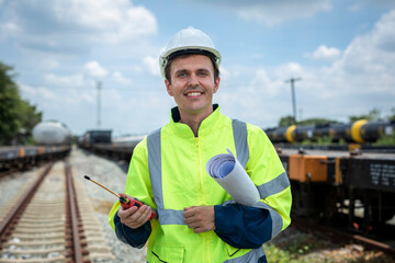 Portrait smiling engineer man with green vest and white hard hat holding walkie-talkie with blueprints working at train station during the day, looking at camera