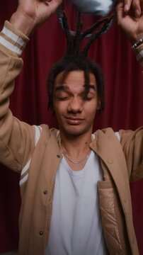 Vertical waist up shot of sleepy-looking young Biracial guy in beige varsity jacket lifting silky eye mask off his face while posing in front of red curtain inside cozy photobooth