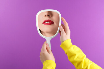 Beautiful woman with red lips looking in handheld mirror on violet background, closeup © New Africa